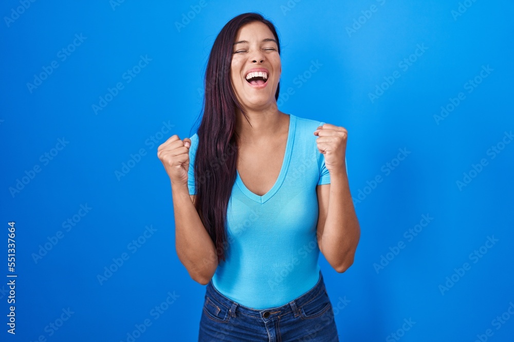 Young hispanic woman standing over blue background excited for success with arms raised and eyes closed celebrating victory smiling. winner concept. Young hispanic woman standing over blue background excited for success with arms raised and eyes closed celebrating victory smiling. winner concept.