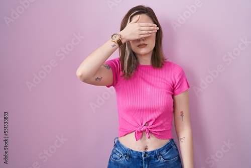 Blonde caucasian woman standing over pink background covering eyes with hand, looking serious and sad. sightless, hiding and rejection concept