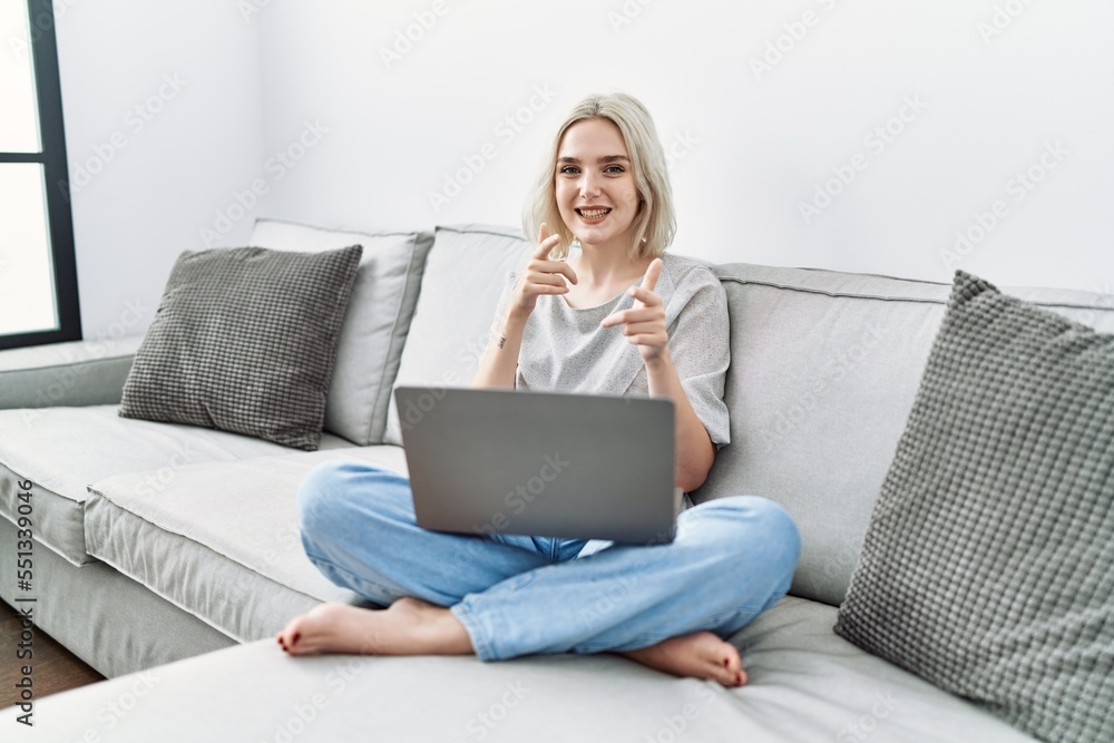 Young caucasian woman using laptop at home sitting on the sofa pointing fingers to camera with happy and funny face. good energy and vibes. Young caucasian woman using laptop at home sitting on the sofa pointing fingers to camera with happy and funny face. good energy and vibes.