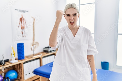 Young caucasian woman working at pain recovery clinic angry and mad raising fist frustrated and furious while shouting with anger. rage and aggressive concept.