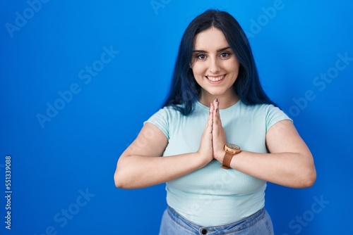 Young modern girl with blue hair standing over blue background praying with hands together asking for forgiveness smiling confident. © Krakenimages.com