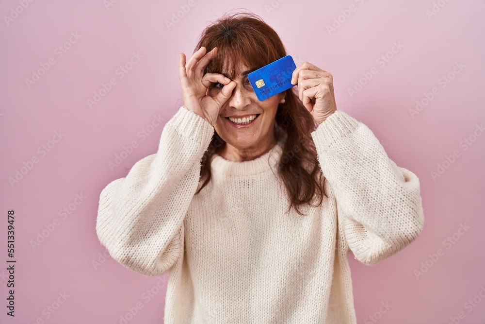 Middle age hispanic woman holding credit card covering eye smiling happy doing ok sign with hand on eye looking through fingers