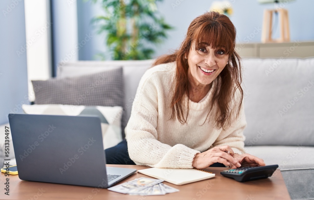 Middle age woman using calculator and laptop sitting on sofa at home Middle age woman using calculator and laptop sitting on sofa at home
