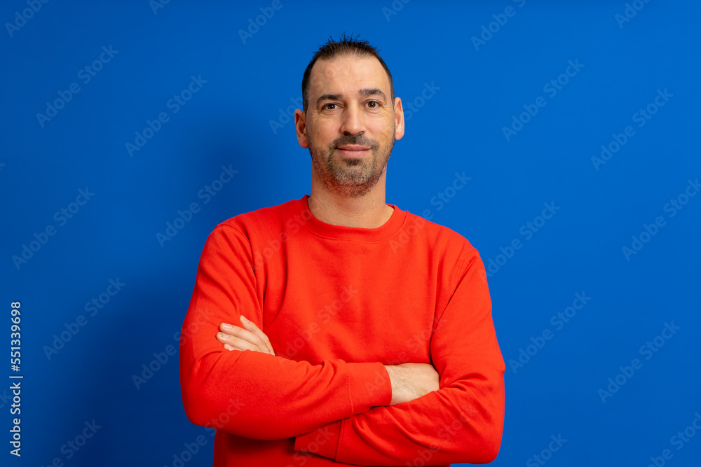 Portrait of smart cheerful attractive bearded hispanic man with arms crossed wearing bright red pullover isolated over blue colored background. Portrait of smart cheerful attractive bearded hispanic man with arms crossed wearing bright red pullover isolated over blue colored background.