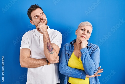 Young brazilian mother and son standing over blue background with hand on chin thinking about question, pensive expression. smiling with thoughtful face. doubt concept.