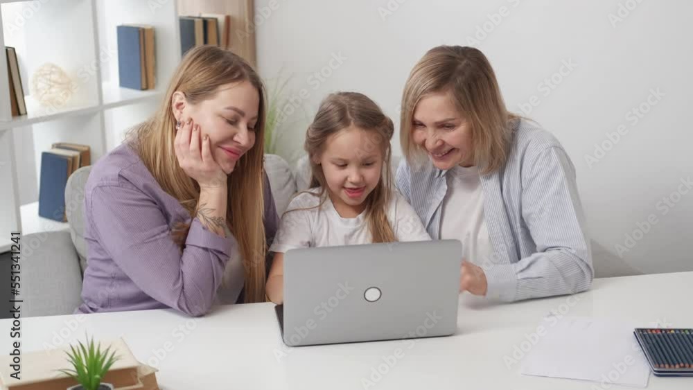 Online game. Kids fun. Close family relationship. Happy girl playing on laptop with supportive mother and grandmother sitting light light room interior. Online game. Kids fun. Close family relationship. Happy girl playing on laptop with supportive mother and grandmother sitting light light room interior.
