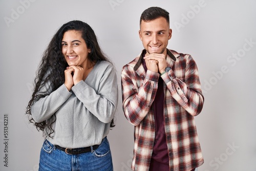 Young hispanic couple standing over white background laughing nervous and excited with hands on chin looking to the side