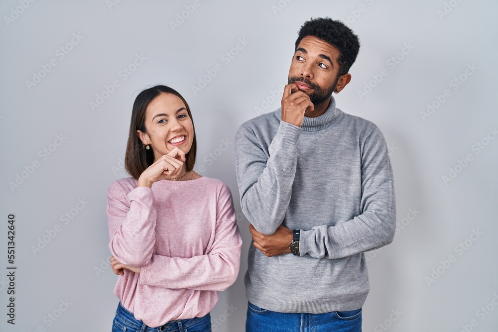 Naklejka premium Young hispanic couple standing together with hand on chin thinking about question, pensive expression. smiling and thoughtful face. doubt concept.