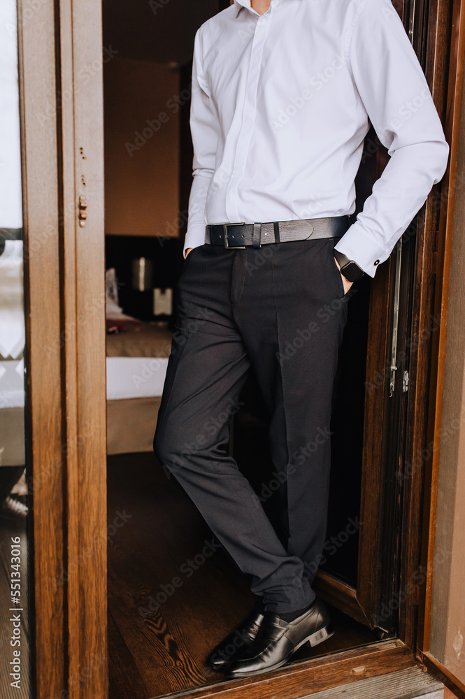 A stylish, rich, fashionable man, businessman, groom stands at the entrance near the wooden door in black pants, leather shoes and a white shirt. Photography, business. A stylish, rich, fashionable man, businessman, groom stands at the entrance near the wooden door in black pants, leather shoes and a white shirt. Photography, business.