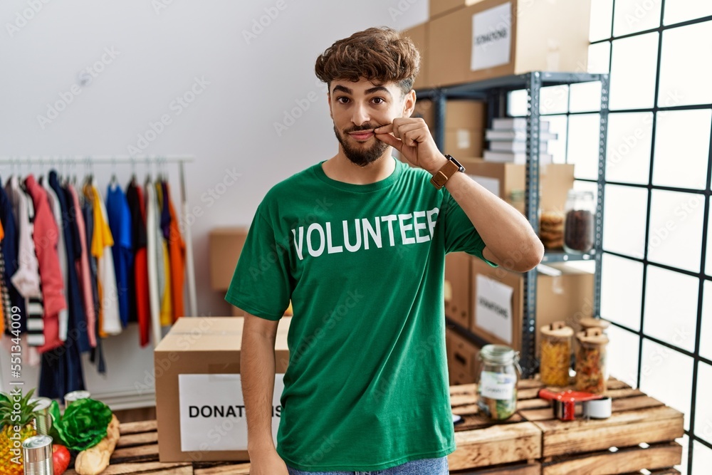 Young arab man wearing volunteer t shirt at donations stand mouth and lips shut as zip with fingers. secret and silent, taboo talking Young arab man wearing volunteer t shirt at donations stand mouth and lips shut as zip with fingers. secret and silent, taboo talking