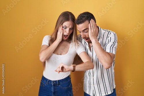 Young couple standing over yellow background looking at the watch time worried, afraid of getting late