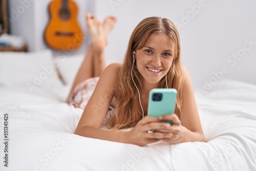 Young blonde girl using smartphone and earphones lying on bed at bedroom