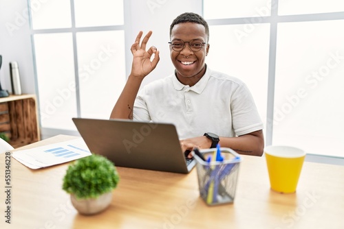 Young african man working at the office using computer laptop smiling positive doing ok sign with hand and fingers. successful expression.