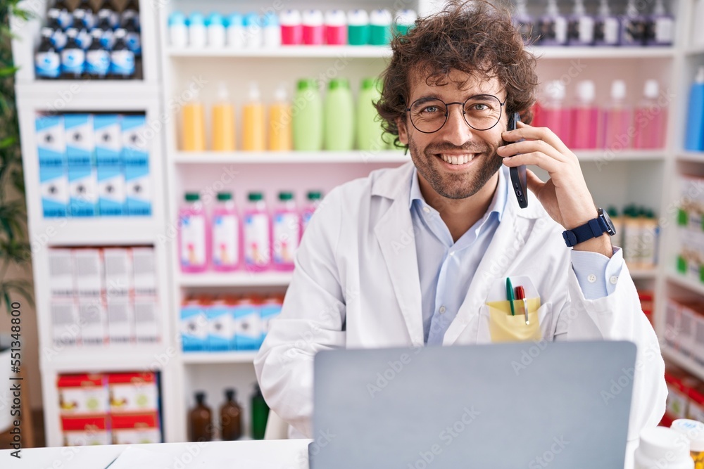 Young hispanic man pharmacist talking on smartphone using laptop at pharmacy Young hispanic man pharmacist talking on smartphone using laptop at pharmacy