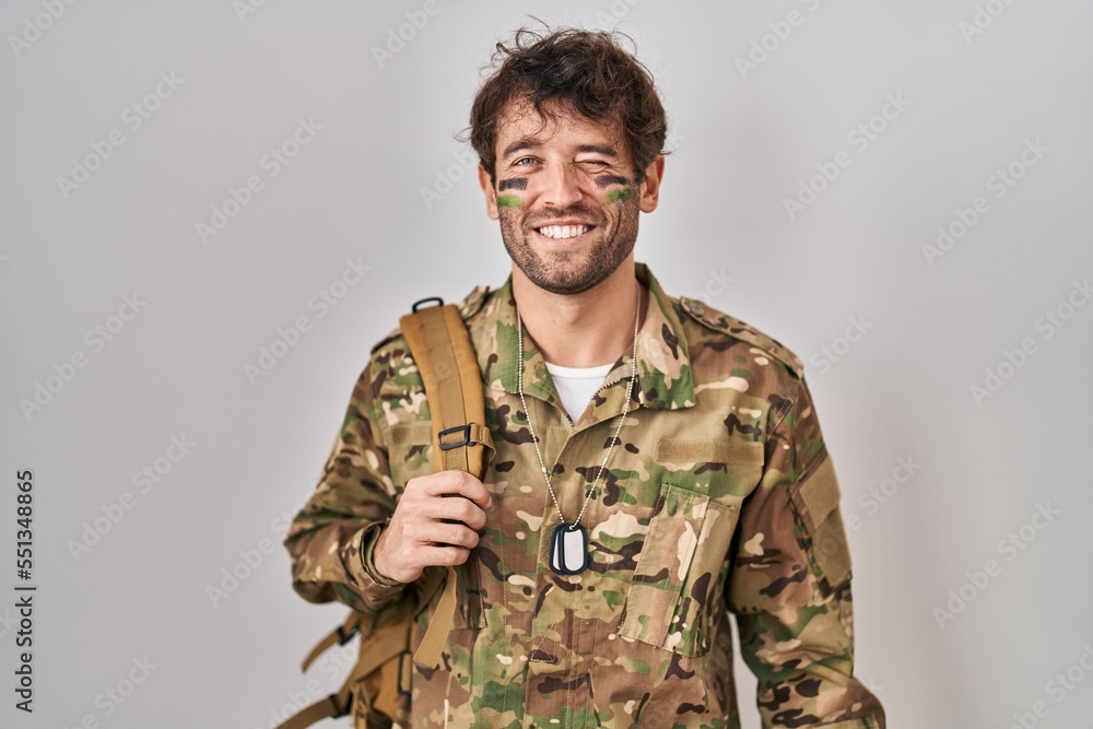 Hispanic young man wearing camouflage army uniform winking looking at the camera with sexy expression, cheerful and happy face.