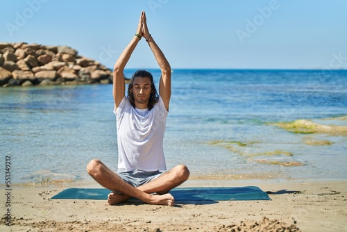 Young hispanic man doing yoga exercise sitting on sand at beach