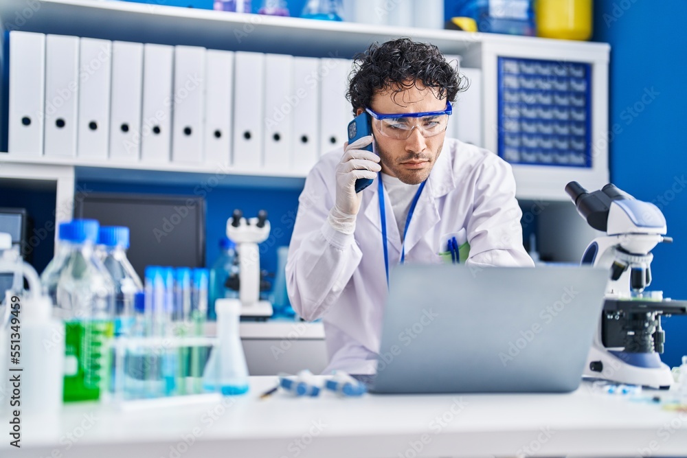 Fototapeta premium Young hispanic man scientist using laptop talking on smartphone at laboratory