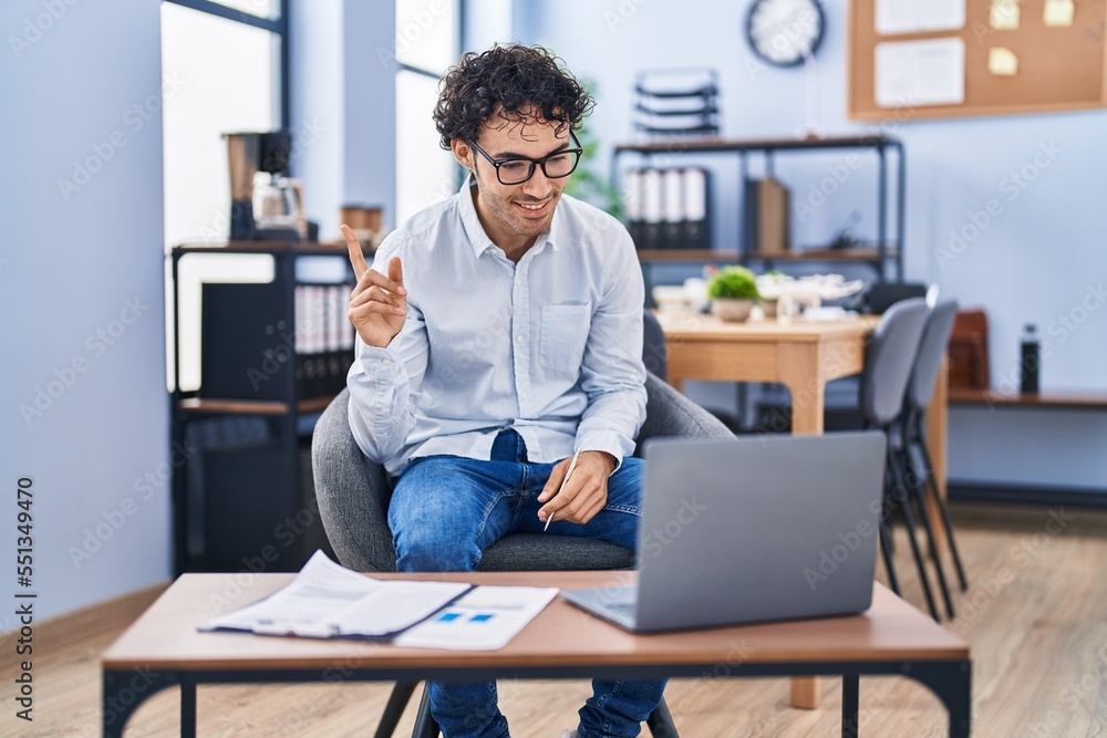 Hispanic man doing video call waving to laptop smiling happy pointing with hand and finger to the side Hispanic man doing video call waving to laptop smiling happy pointing with hand and finger to the side