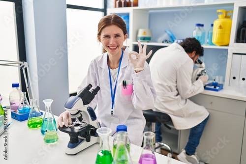Young two people working at scientist laboratory doing ok sign with fingers  smiling friendly gesturing excellent symbol