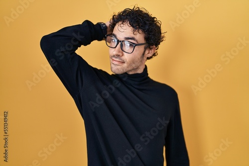 Hispanic man standing over yellow background smiling confident touching hair with hand up gesture, posing attractive and fashionable