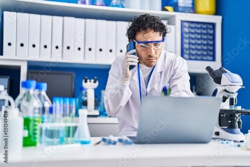 Young hispanic man scientist using laptop talking on smartphone at laboratory