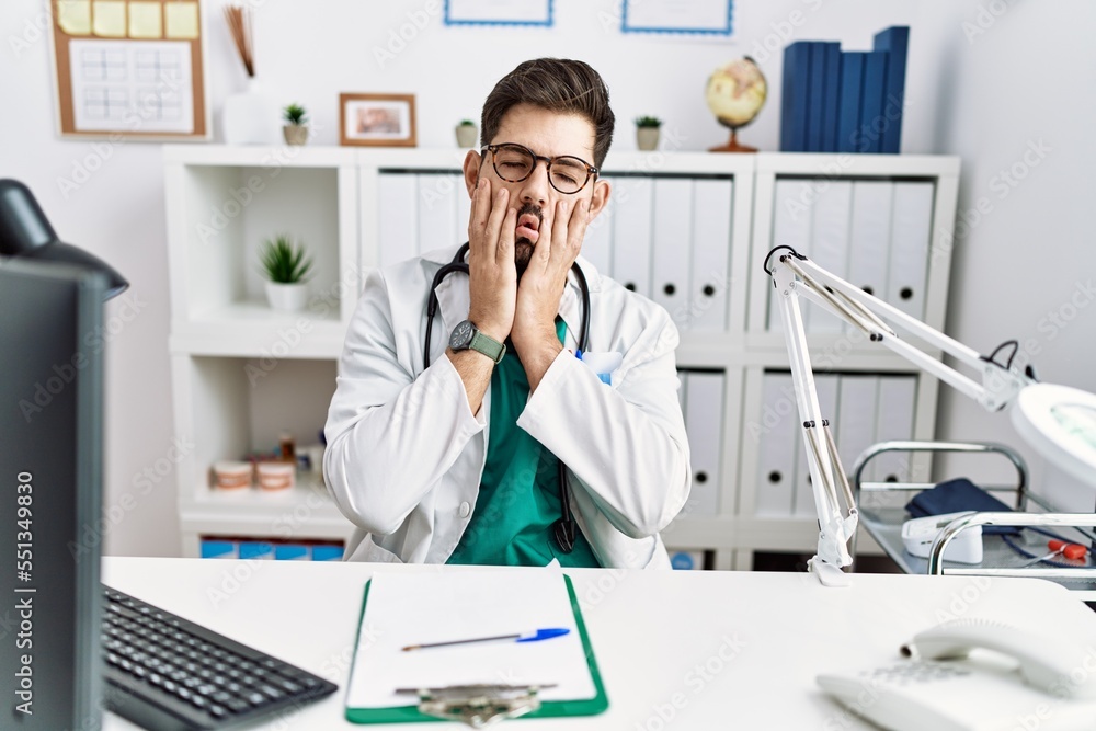 Young man with beard wearing doctor uniform and stethoscope at the clinic tired hands covering face, depression and sadness, upset and irritated for problem Young man with beard wearing doctor uniform and stethoscope at the clinic tired hands covering face, depression and sadness, upset and irritated for problem