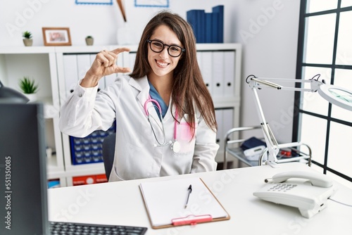 Young doctor woman wearing doctor uniform and stethoscope at the clinic smiling and confident gesturing with hand doing small size sign with fingers looking and the camera. measure concept.