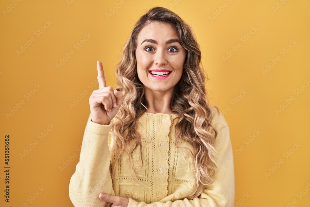 Young caucasian woman standing over yellow background pointing finger up with successful idea. exited and happy. number one. Young caucasian woman standing over yellow background pointing finger up with successful idea. exited and happy. number one.