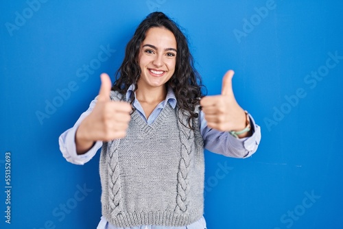 Young brunette woman standing over blue background approving doing positive gesture with hand, thumbs up smiling and happy for success. winner gesture.