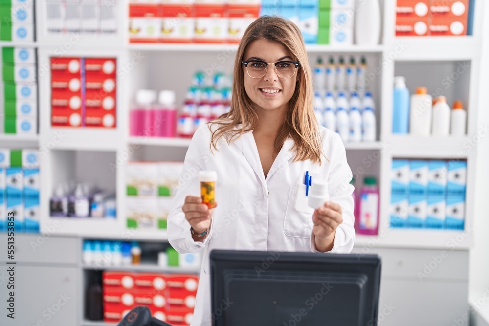 Young woman pharmacist smiling confident holding pills bottles at pharmacy