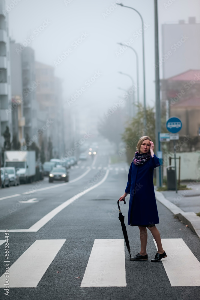 A woman with an umbrella at a crosswalk in overcast weather. A woman with an umbrella at a crosswalk in overcast weather.