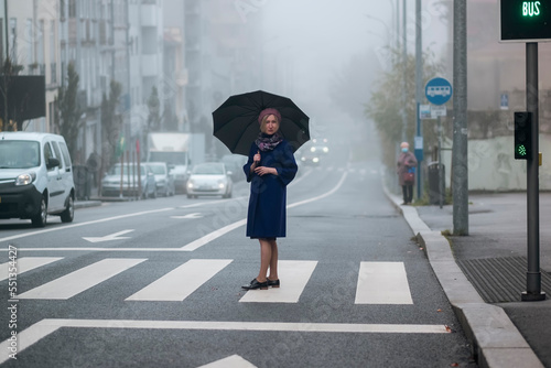 A woman in an overcoat with an umbrella stands by the road in the fog. photo