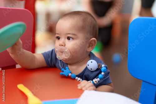 Adorable chinese toddler using pacifier holding dish toy at kindergarten