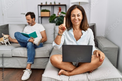 Hispanic middle age couple at home, woman using laptop showing and pointing up with finger number one while smiling confident and happy.