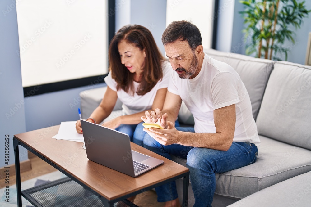 Obraz premium Middle age man and woman couple accounting using smartphone and laptop at home