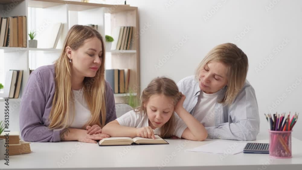 Study reading. Curious girl. Supportive family. Mother and grandmother helping cute kid study new book sitting desk in light room interior. Study reading. Curious girl. Supportive family. Mother and grandmother helping cute kid study new book sitting desk in light room interior.