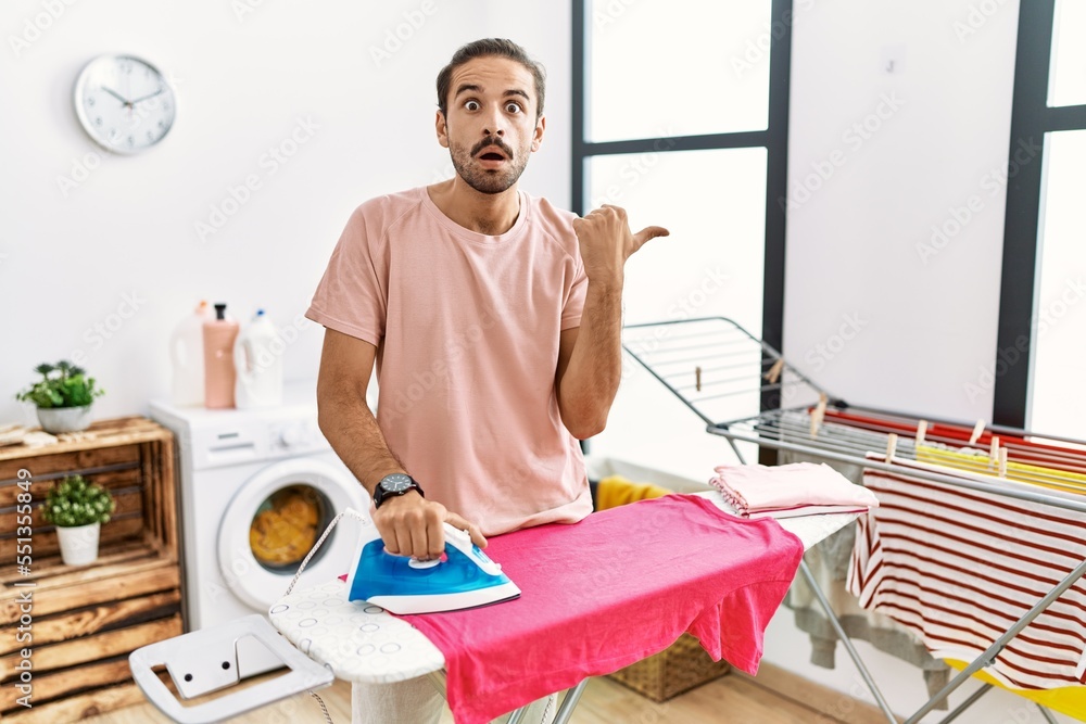 Young hispanic man ironing clothes at home surprised pointing with hand finger to the side, open mouth amazed expression. Young hispanic man ironing clothes at home surprised pointing with hand finger to the side, open mouth amazed expression.