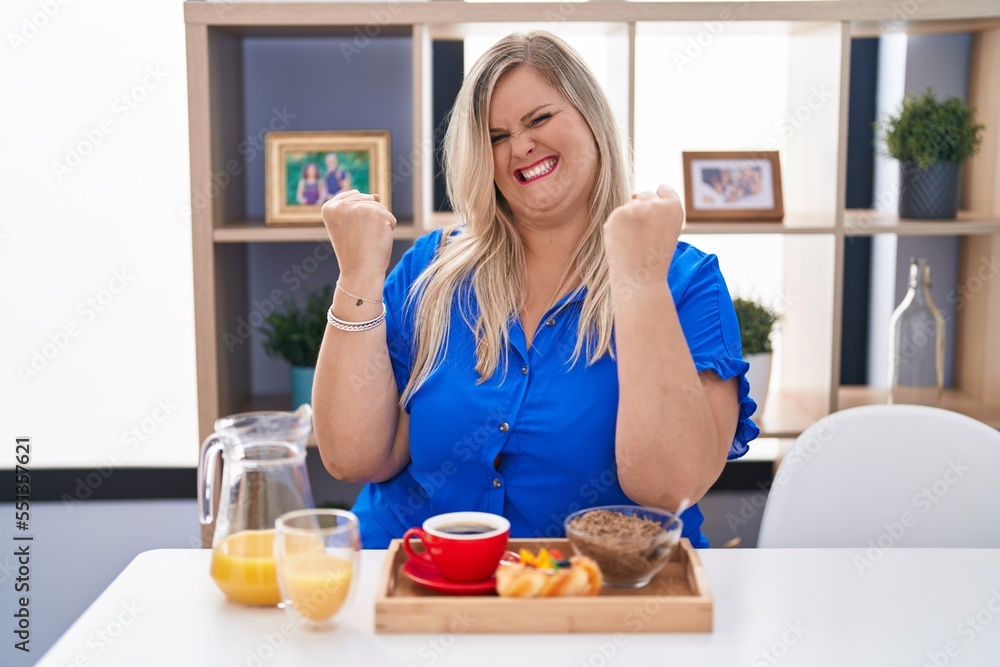 Caucasian plus size woman eating breakfast at home very happy and excited doing winner gesture with arms raised, smiling and screaming for success. celebration concept. Caucasian plus size woman eating breakfast at home very happy and excited doing winner gesture with arms raised, smiling and screaming for success. celebration concept.