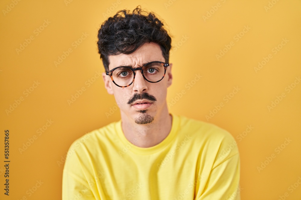 Hispanic man wearing glasses standing over yellow background skeptic and nervous, disapproving expression on face with crossed arms. negative person. Hispanic man wearing glasses standing over yellow background skeptic and nervous, disapproving expression on face with crossed arms. negative person.