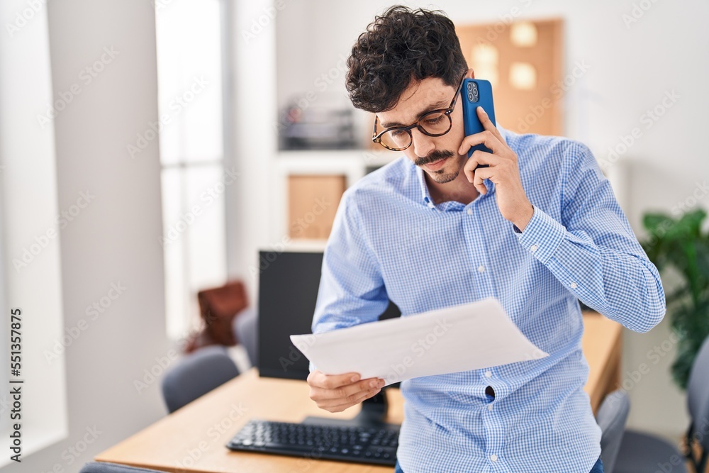 Obraz premium Young caucasian man business worker talking on smartphone reading document at office