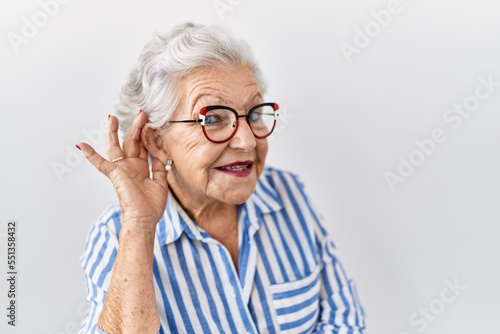 Senior woman with grey hair standing over white background smiling with hand over ear listening an hearing to rumor or gossip. deafness concept.