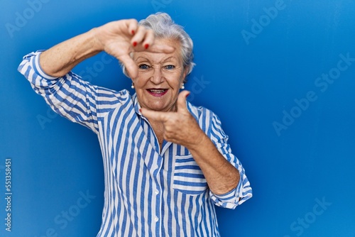 Senior woman with grey hair standing over blue background smiling making frame with hands and fingers with happy face. creativity and photography concept.
