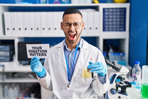 Young hispanic man working at scientist laboratory holding your donation matters holding blood sample angry and mad screaming frustrated and furious, shouting with anger. rage and aggressive concept.