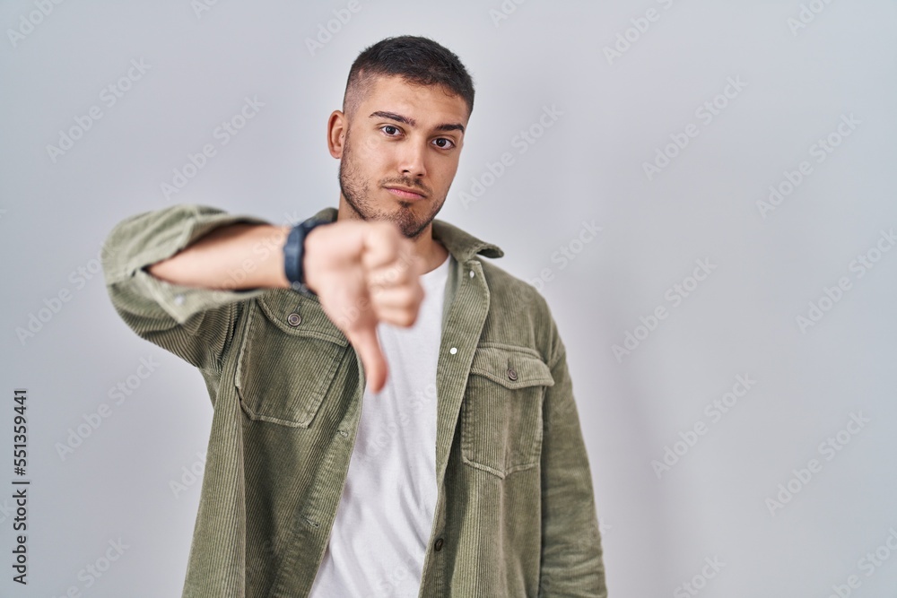 Young hispanic man standing over isolated background looking unhappy and angry showing rejection and negative with thumbs down gesture. bad expression. Young hispanic man standing over isolated background looking unhappy and angry showing rejection and negative with thumbs down gesture. bad expression.