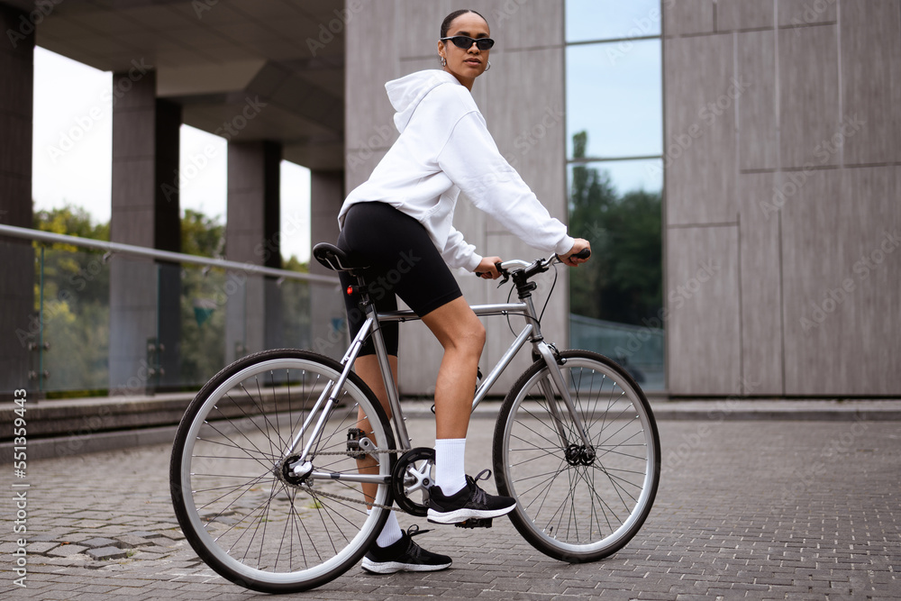 Trendy african american woman in sunglasses standing near bike on street at daytime Trendy african american woman in sunglasses standing near bike on street at daytime
