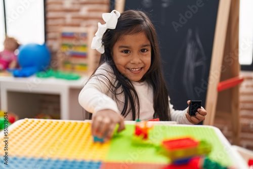 Adorable hispanic girl playing with construction blocks sitting on table at kindergarten