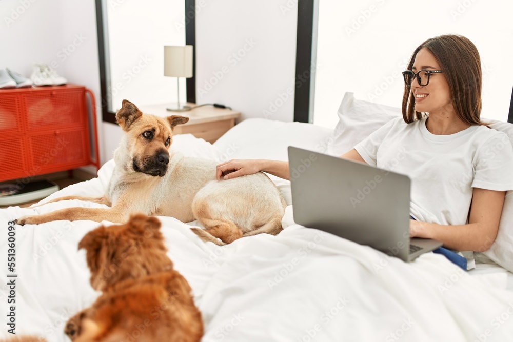 Young hispanic woman using laptop sitting on bed with dogs at bedroom