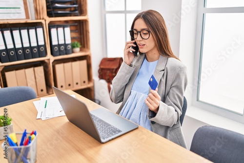 Young beautiful hispanic woman business worker talking on smartphone holding credit card at office
