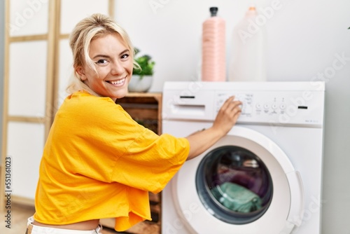 Young blonde girl doing laundry putting clothes into washing machine at home.