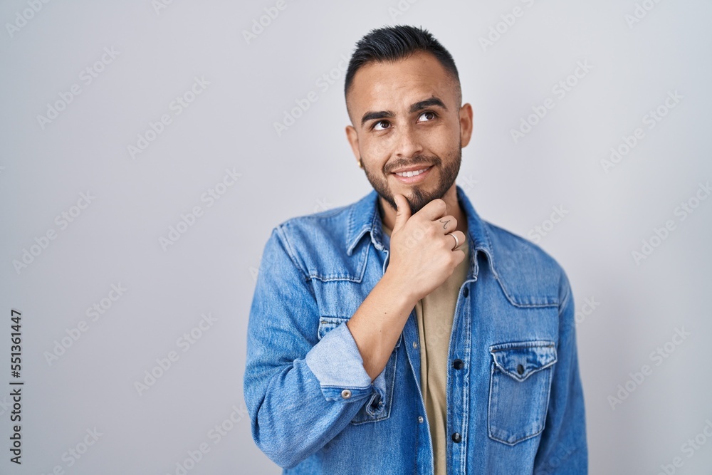 Young hispanic man standing over isolated background with hand on chin thinking about question, pensive expression. smiling and thoughtful face. doubt concept. Young hispanic man standing over isolated background with hand on chin thinking about question, pensive expression. smiling and thoughtful face. doubt concept.